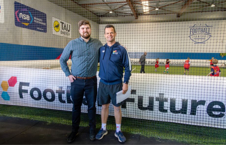 Two men standing at a football training facility, supporting Rise's PWD Employment program, with children playing in the background.